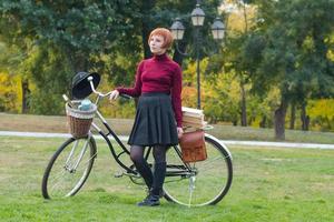 mujer joven en el parque de otoño lee un libro, hermosa mujer pelirroja con bicicleta en la hierba verde foto