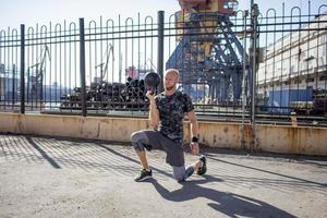 Young bearded male athlete training in industrial zone in sunny day, kettlebells exercises outdoors, urban background photo