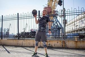 Young bearded male athlete training in industrial zone in sunny day, kettlebells exercises outdoors, urban background photo