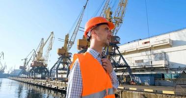 male worker of sea harbor in orange helmet and safety west, cranes and sea background photo