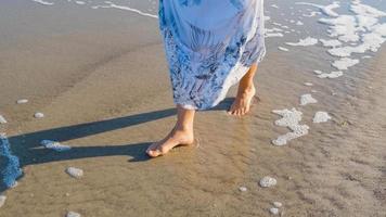 joven hermosa mujer vestida con un vestido blanco camina descalzo en la playa de verano foto