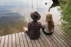 Young couple of travelers sitting on the wooden pier near river, two hipsters with hats and dreadlocks on vacation photo