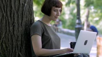 jeune femme travaille avec un ordinateur portable à l'extérieur dans le parc d'été, femme avec ordinateur sur l'herbe verte 4k shot video