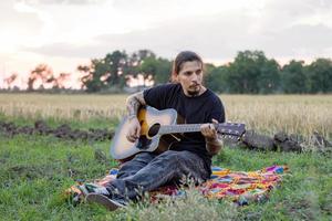 Young tattooed male play on acoustic guitar in summer fields during beautiful sunset photo