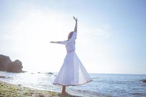 mujer joven caminando en la playa de la mañana en un hermoso vestido blanco. mujer en forma pasando un buen rato durante el amanecer. foto