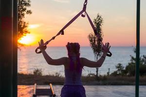 Beautiful fit woman in pink and purple sportwear training on outdoor gym in morning, exercises with suspension straps in park photo