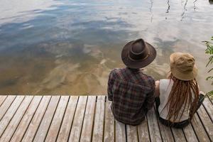 Young couple of travelers sitting on the wooden pier near river, two hipsters with hats and dreadlocks on vacation photo