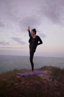 Fit woman doing yoga stretching exercise outdoor in beautiful mountains landscape. Female on the rock with sea and sunrise or sunset background training asans. Silhouette of woman in yoga poses photo