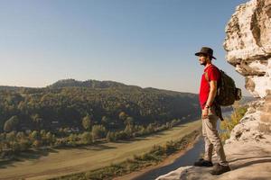 Young bearded male traveler in hat stand on the cliff in sunny day photo