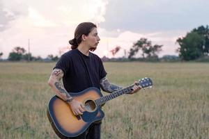 Young tattooed male play on acoustic guitar in summer fields during beautiful sunset photo