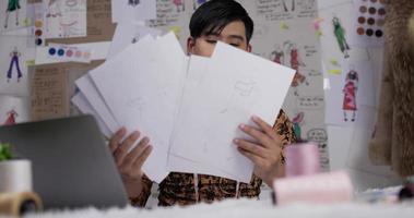 Aziatische mannelijke modeontwerper die gegevens op papier controleert of tekeningen overweegt terwijl hij aan dek zit. gerichte man kledingontwerper aan het werk in zijn kantoor.