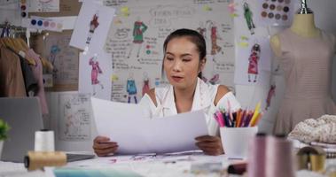 asiatische modedesignerin, die farbmuster durchsieht oder daten auf papier überprüft und zeichnungen an bord betrachtet. Fokussierte Modedesignerin, die arbeitet, während sie in ihrem Büro am Schreibtisch sitzt. video