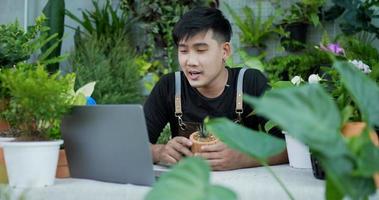 retrato de un joven jardinero asiático feliz que vende en línea en las redes sociales a través de una computadora portátil en el jardín. hombre en videollamada de máscara facial en computadora portátil. vegetación doméstica, venta en línea y concepto de hobby video