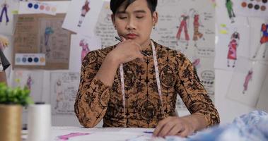 retrato de homem asiático estilista cansado desenha uma roupa de esboço enquanto está sentado no estúdio. startup pequeno empresário está em processo de criação de uma nova coleção de roupas. video