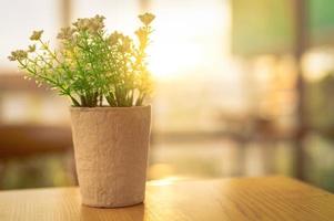 Fake white flowers with green leaves in recycled paper pot on brown wooden table in coffee cafe in the morning with sunshine. Plastic flower in pot. Home office interior and living room decor concept. photo