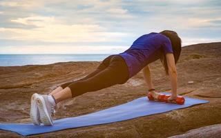 Asian woman workout in the morning at stone beach. Fit woman hand hold red dumbbell and push up on blue yoga mat for body strength. Weight training exercise for healthy lifestyle. Flat butt woman. photo