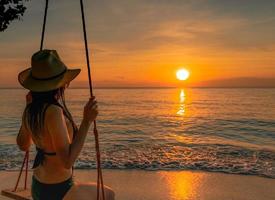 Sexy woman wear bikini and straw hat swing the swings at tropical beach on summer vacation at sunset. Girl in swimwear sit on swings and watching beautiful sunset. Summer vibes. Woman travel alone.
