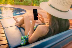 Asian woman with hat and swimsuit sitting in chair at poolside and using smartphone on summer vacation. Luxury life. Checking News on mobile phone application by swimming pool. Girl with nail manicure photo