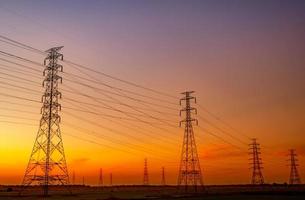 pilón eléctrico de alto voltaje y cable eléctrico con cielo al atardecer. postes de electricidad concepto de potencia y energía. torre de red de alto voltaje con cable de alambre en el campo de cultivo de arroz cerca de la fábrica industrial. foto