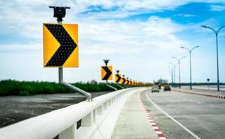 Black and yellow arrow on curve traffic sign on the bridge with solar cell panel ob blurred background of concrete road and car near mud flat and mangrove forest with beautiful blue sky and clouds. photo