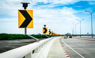 Black and yellow arrow on curve traffic sign on the bridge with solar cell panel ob blurred background of concrete road and car near mud flat and mangrove forest with beautiful blue sky and clouds. photo