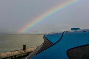 enfoque selectivo en el maletero del camión suv de lujo azul con gotas de lluvia en el arco iris y el fondo del cielo tormentoso. coche aparcado en el aparcamiento al aire libre junto a la playa del mar en un día lluvioso. coche mojado con gota de agua. foto