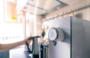 Water purifier in apartment kitchen on blurred background of woman hand holding handle of modern electric kettle. Selective focus on on and off button of water purifier machine. Water filter system. photo