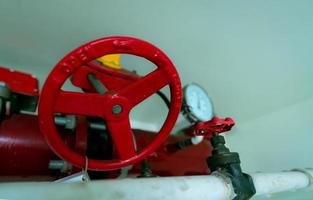 Red hand wheel of main supply water piping in the fire extinguishing system and white water supply pipe. Fire sprinkler system with red pipes hanging from ceiling inside building. Fire Suppression. photo