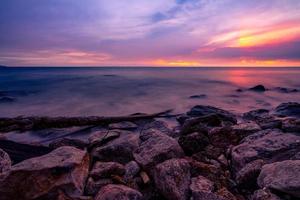 Stone beach and calm sea at dusk with purple sunset sky. Tropical sea. Skyline in the evening with golden, purple, and red sky. Seascape. Summer vacation travel. Rock coast. Beauty in ocean nature. photo