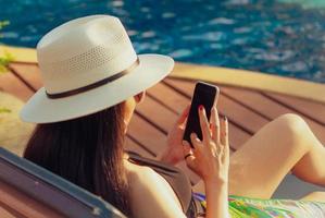 Asian woman with hat and swimsuit sitting in chair at poolside and using smartphone on summer vacation. Luxury life. Checking News on mobile phone application by swimming pool. Girl with nail manicure photo