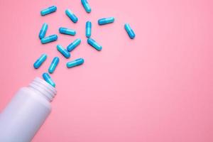 Blue capsule pills spread out of white plastic drug bottle on pink background. Pharmacy banner. Online pharmacy. Painkiller medicine and antibiotic drug resistance concept. Pharmaceutical industry. photo