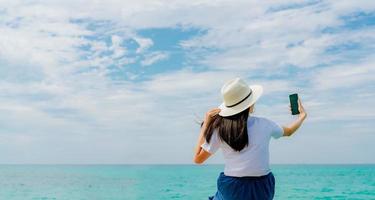 Young Asian woman wear straw hat use smartphone taking selfie at wooden pier. Summer vacation at tropical paradise beach. Happy girl travel on holiday. Woman enjoy and relax life. Summer vibes. photo