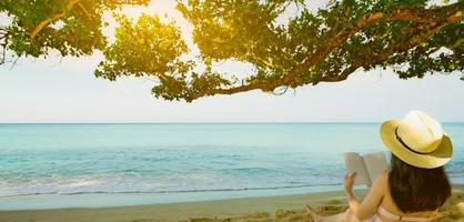 Women sit and reading a book under the tree at seaside. Back view of sexy Asian woman with straw hat relaxing and enjoying holiday at tropical paradise sand beach. Summer vacation. Summer vibes. photo