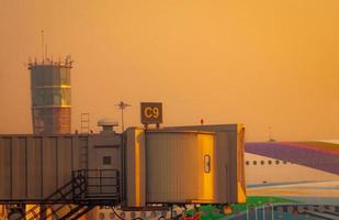 avión comercial estacionado en el puente de reacción para el despegue de pasajeros en el aeropuerto. puente de embarque de pasajeros de aviones acoplado con cielo dorado al atardecer cerca de la torre de control de tráfico aéreo en el aeropuerto. foto