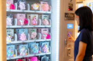 desenfoque mujer asiática presionando el botón en la máquina expendedora para comprar muñeca. mujeres turistas comprando muñecas en máquinas expendedoras automáticas en el centro comercial del aeropuerto. dispositivo de autoservicio. consumidor y máquina expendedora. foto