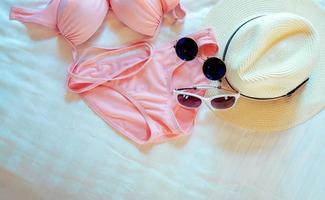 Top view of bikini swimsuit, sunglasses, and straw hat on bed sheet. Woman's swimwear and beach accessories on bed of resort. Pink bikini swimwear set for the beach. Summer vacation and summer vibes. photo