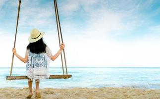 Asian woman in casual style wear hat and sandals sit on swings at sand beach  and looking beautiful tropical paradise sea and sky. Summer vacation. Summer vibes. Enjoying and relaxing girl on holiday. photo