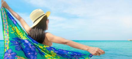 Back view of Asian woman wear swimsuit and opened arms at tropical beach on sunny day with beautiful blue sky and white clouds. Woman travel on summer vacation. Summer vibes. Happy girl. photo