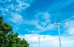 Horizontal axis wind turbine with blue sky and white clouds near green tree. Wind energy in eco wind farm. Green energy concept. Renewal energy. Alternative electricity source. Sustainable resources. photo