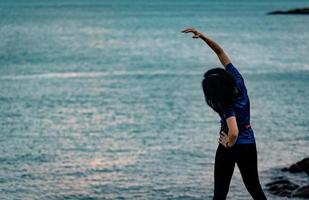 entrenamiento de mujeres asiáticas por la mañana en la playa de piedra con un hermoso cielo de amanecer. Corredor de mujer en forma estirando el cuerpo antes de correr. ejercicio cardiovascular para un estilo de vida saludable. chica activa entrenando sola. foto