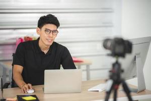 online sale and social media marketing concept, young Asian man working with camera to live streaming to sale a product and showing packet to review, broadcast cyberspace and blog shop photo