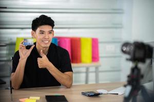 online sale and social media marketing concept, young Asian man working with camera to live streaming to sale a product and showing packet to review, broadcast cyberspace and blog shop