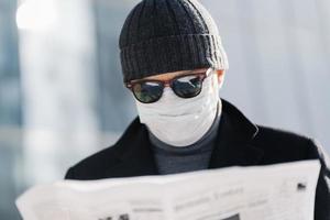 Photo of attentive man wears blak sunglasses, hat and medical mask on face during quarantine and coronavirus outbreak, reads press, finds out news from newspaper, stands against blurred background