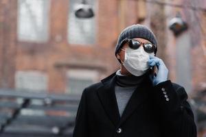 Sick man wears protective mask on face during virus pandemic, has telephone conversation with friend, wears black coat and shades, poses outdoor. Protect your health. Coronavirus, panic concept photo