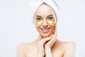 Pretty smiling lady has towel on head touches cheeks, applies golden hydrogel patches, stands shirtless indoor, removes wrinkles, isolated on white wall, has healthy fresh facial skin. High resolution