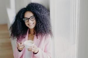 Photo of pleasant looking curly haired lady with toothy smile, dressed formally, poses indoor, holds mug of latte, has coffee break after long hours office work, looks through eyewear, feels happy