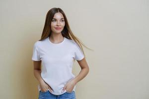 Thoughtful brunette woman looks aside, wears casual white t shirt, keeps hands in pockets, looks aside, isolated over beige background, has relaxed face expression, empty space for your promotion photo