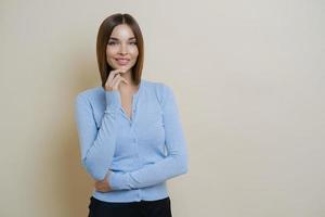 Half length shot of pretty slim woman dressed in casual blue jumper, keeps hand under chin, enjoys smooth perfect healthy skin, ready for work, isolated over beige background, blank space aside photo