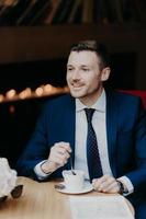 Vertical shot of handsome male in formal clothing, has cheerful expression, has dinner break at cafeteria, drinks aromatic coffee, looks happily somewhere, thinks about something. Business and rest photo
