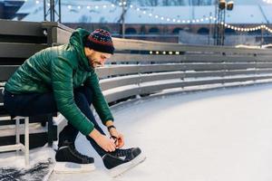 Photo of skilled male model with happy expression laces up skates, dressed in green anorak, sits on ice ring, going to be involved in his favourite hobby. Man has fun and entertainment outdoor